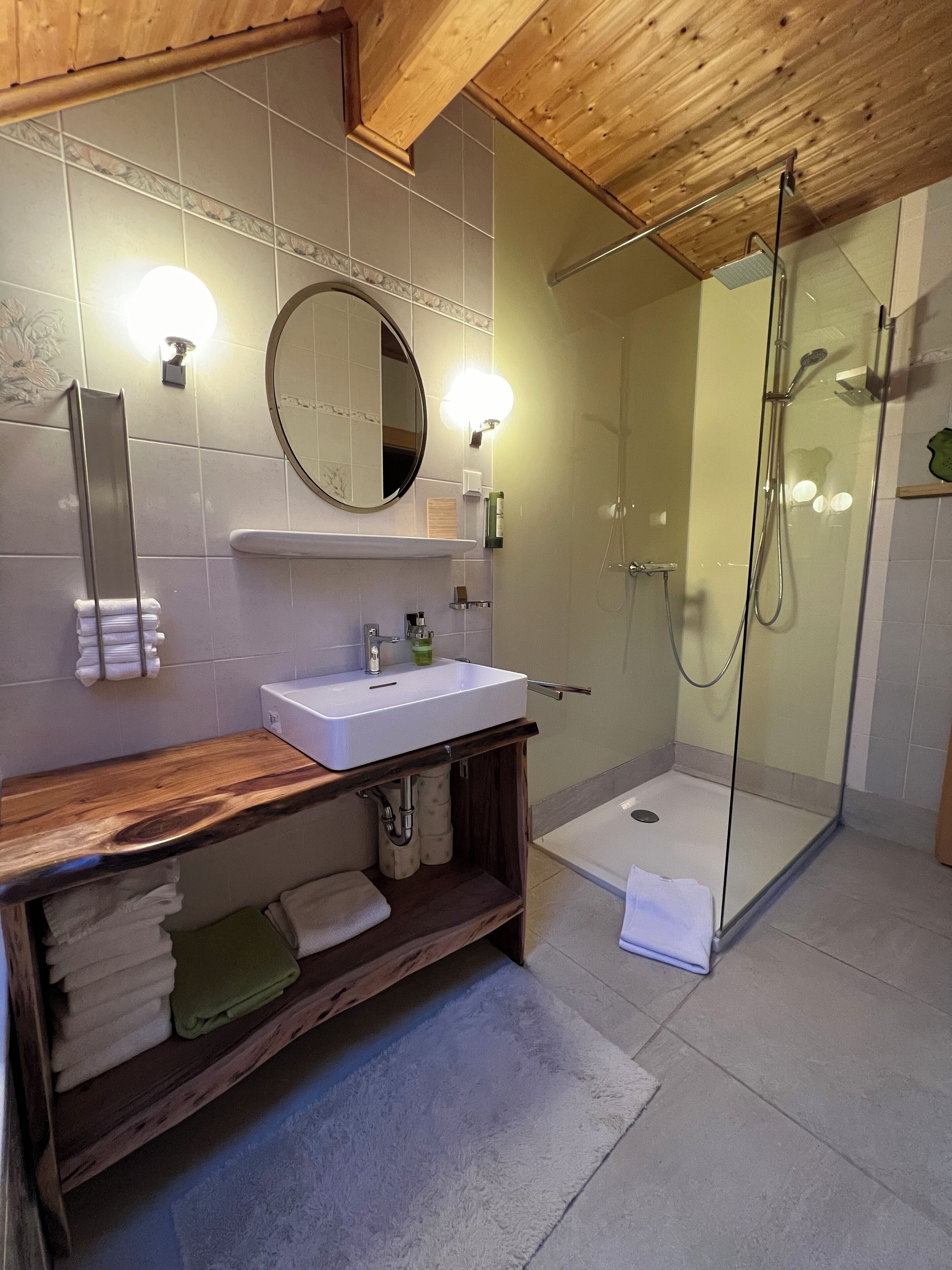 Un baño moderno con tocador de madera, espejo redondo, ducha con mampara de vidrio y suelo de baldosas.