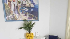 A room with a white console table, a potted plant, a painting of a coastal town, and a gray chair.