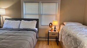 Two comfortable beds and a nightstand are situated in this serene bedroom, with a window and lamp adding to the cozy ambiance.