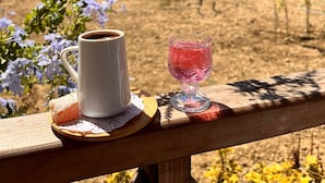 A wooden deck with a white mug and a pink glass on a coaster, surrounded by purple flowers.