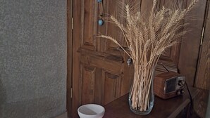 A wooden table with a white ceramic vase containing dried plants, a small radio, and a lit candle on a coiled mat.
