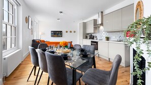 A modern kitchen with white cabinets, a dining area with a glass table and grey chairs, and a cozy living room with a sofa and orange cushions.
