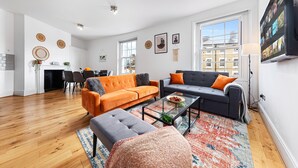 A modern living room with a sofa, ottoman, and coffee table, a dining area with chairs, and a kitchen in the background.