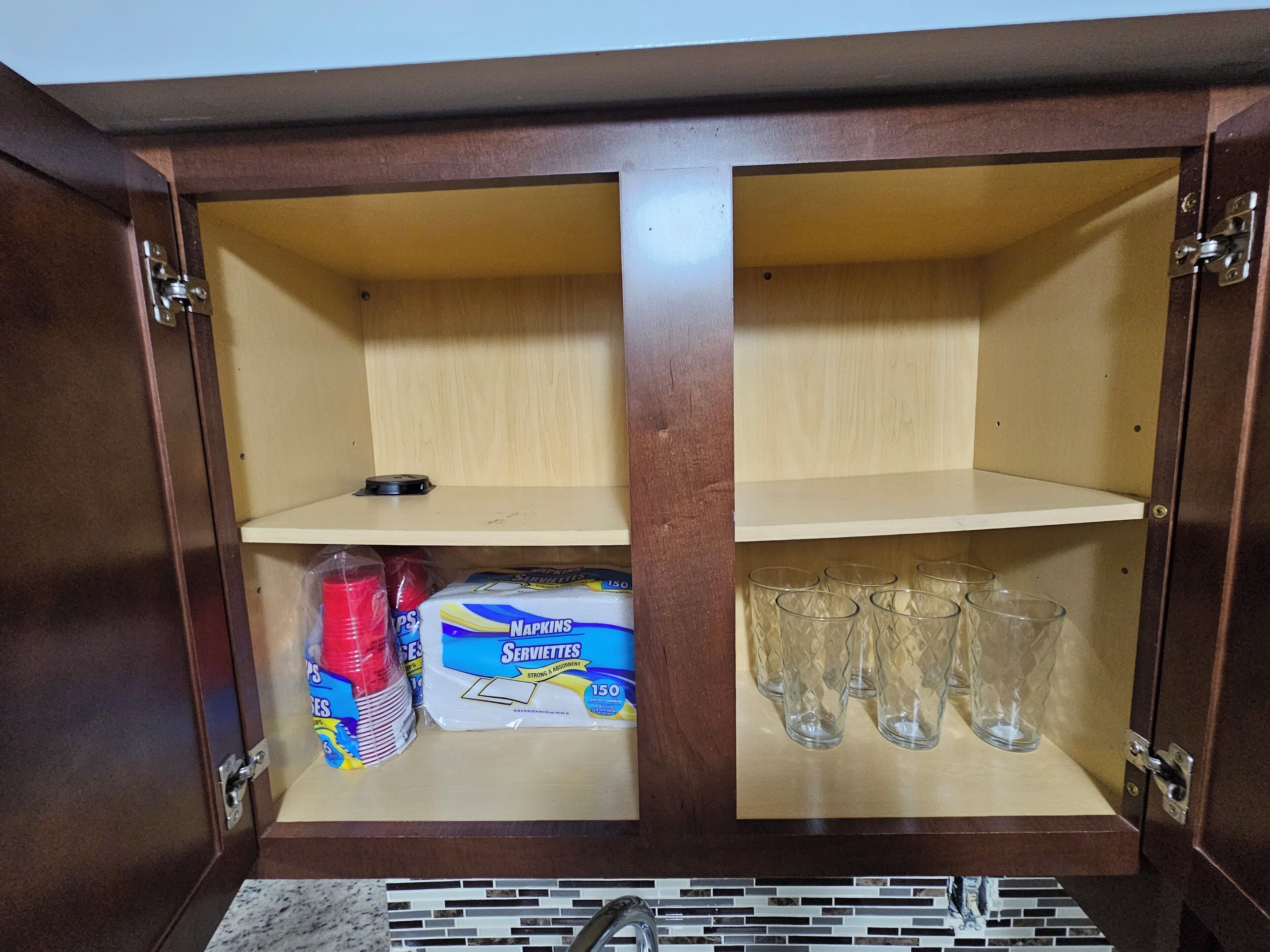 Un meuble en bois avec deux étagères ; l’une contient une boîte de serviettes en papier et des gobelets rouges, et l’autre est garnie de plusieurs verres transparents.