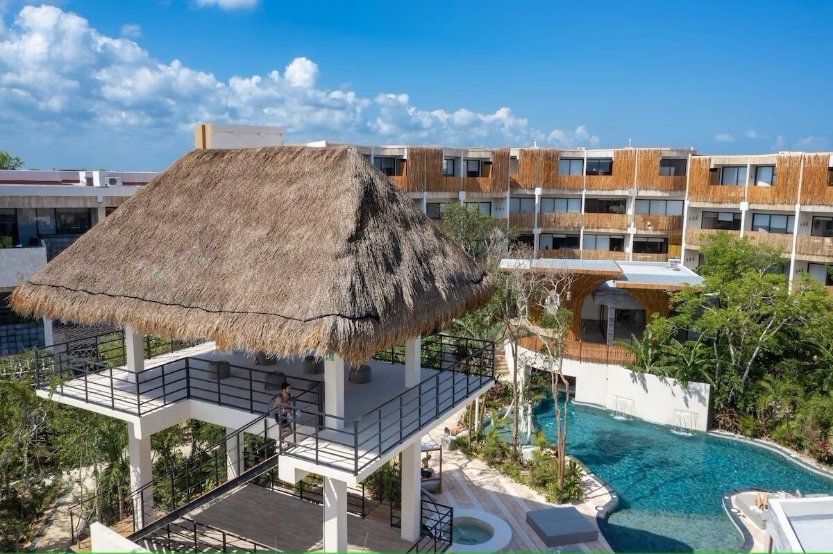 Un complexe hôtelier doté d’une structure à toit de chaume, d’une piscine et de bâtiments modernes avec balcons.