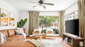 Una sala de estar con un sofá de cuero marrón, dos sillones, una mesa de centro, un televisor sobre un mueble de madera y un ventanal con cortinas.