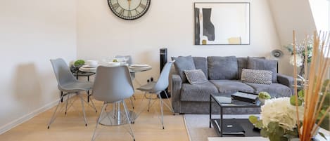 Un salon moderne avec un coin repas, comprenant une table ronde, des chaises grises, un canapé gris et une grande horloge murale.