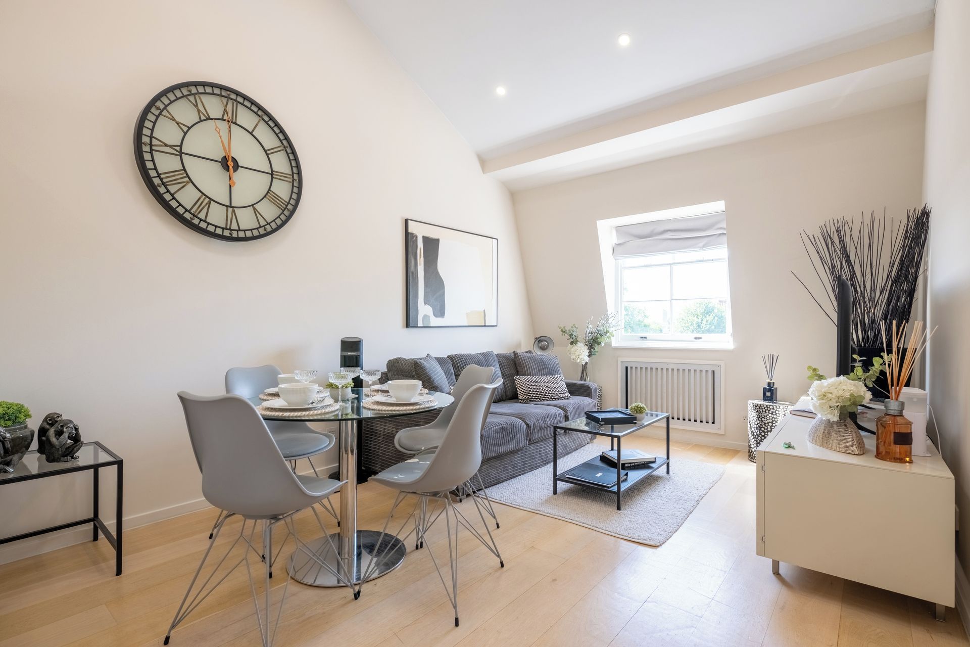 A modern living room with a dining area, a large wall clock, and a window with natural light.
