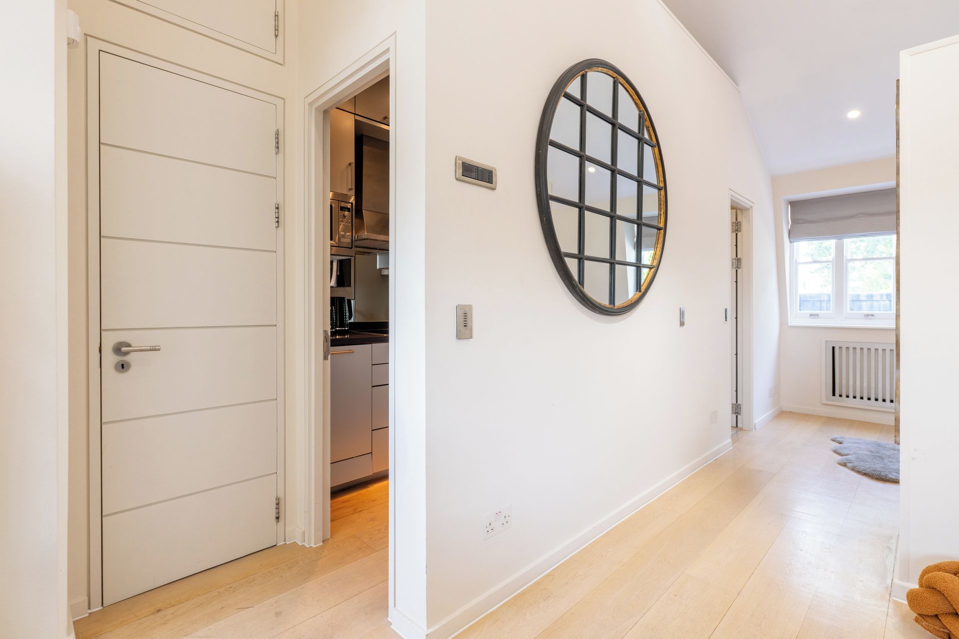 A hallway with a white door, a kitchen area, and a large circular mirror on the wall.