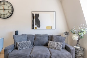 Una sala de estar moderna con un sofá gris, un reloj, un cuadro abstracto enmarco y un florero con flores blancas.