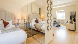 A modern bedroom with a large mirror, a bed with multiple pillows, bedside tables with lamps, and a view of a living area with a sofa and a window. - Newly Renovated 1BDR in Knightsbridge with WiFi (London)