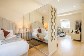 A modern bedroom with a large mirror, a bed with multiple pillows, bedside tables with lamps, and a view of a living area with a sofa and a window. - Newly Renovated 1BDR in Knightsbridge with WiFi (London)
