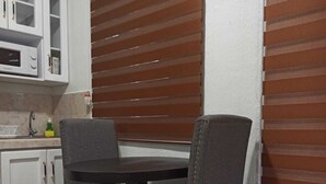 A dining area with a table and four chairs, a kitchen with white cabinets, and a window with brown blinds. - Silvia House II Acropoli Sonsonate. (Sonsonate)