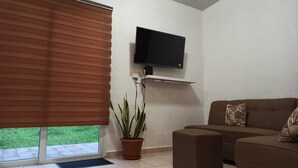 A living room with a flat-screen TV mounted on the wall, a brown sofa with patterned cushions, a potted plant, and a sliding glass door leading to a garden. - Silvia House II Acropoli Sonsonate. (Sonsonate)