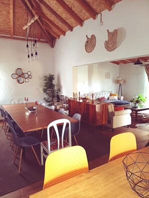 Een ruime eethoek met een houten tafel en stoelen, een keuken op de achtergrond en een hoog plafond met zichtbare balken.
