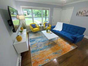A modern living room with a blue couch, yellow armchairs, a white coffee table, and a large window with a view of a green landscape.