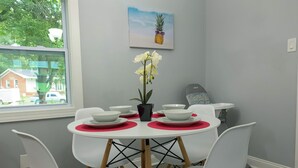 Een eetgedeelte met een ronde tafel, witte stoelen en rode placematen. Er is een raam met uitzicht op huizen en bomen.