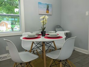 A dining area with a round table, white chairs, and red placemats. There is a window with a view of houses and trees.