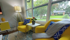 Een gele bank en fauteuil in een kamer met een groot raam, een tafeltje en een lamp.