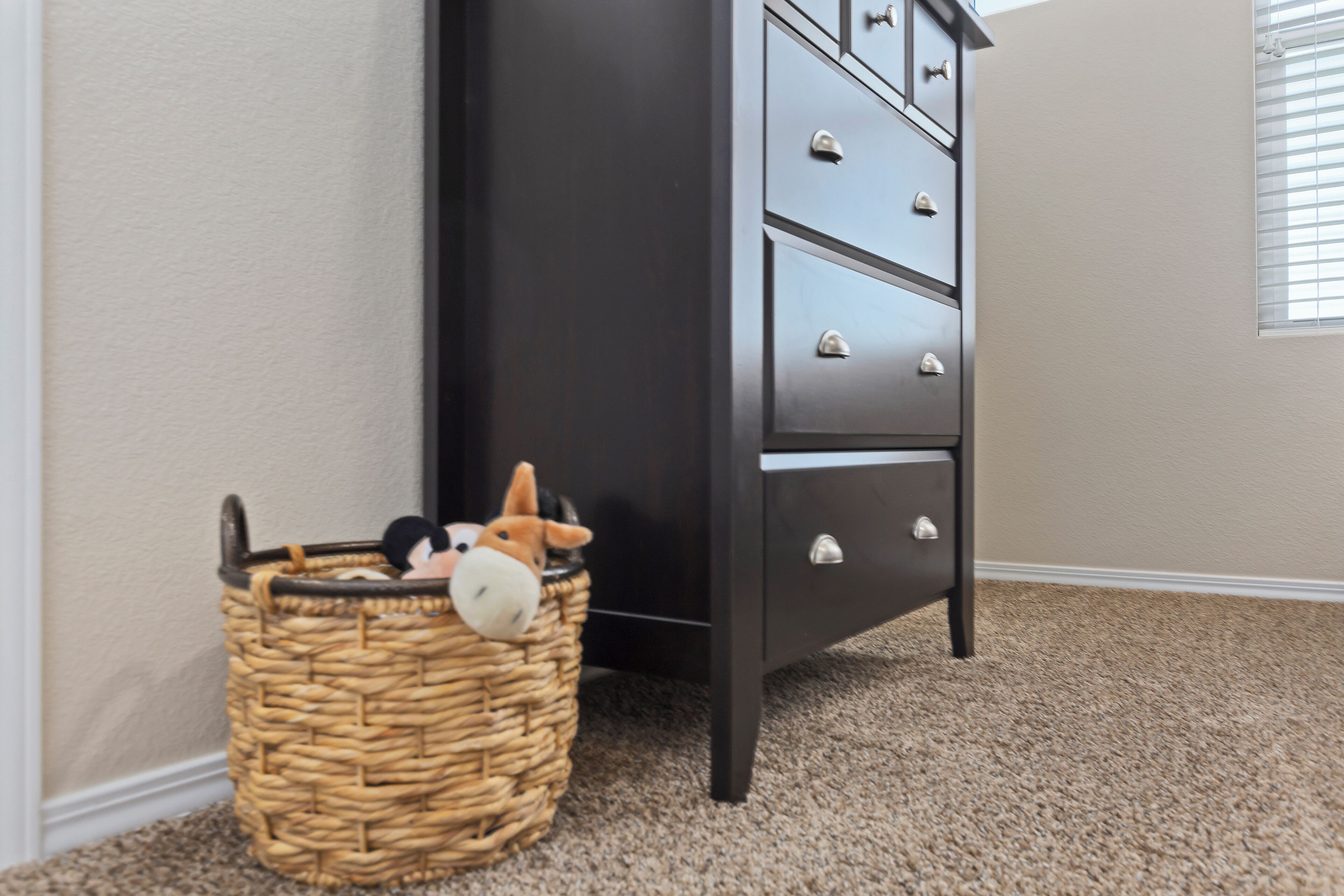 A corner of a room with a dark-colored dresser, a wicker basket with stuffed toys, and a carpeted floor.