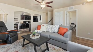 A living room with a grey sofa, black armchair, and a coffee table. There is a ceiling fan, a kitchen in the background, and a staircase leading to another level. - Sleeps 9, Pet Friendly, Workspace, Easy Access to Dining and Shops (Meridian)