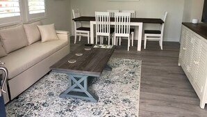 A cozy living room with a sofa, a wooden coffee table, and a dining area with white chairs and a table. - The Mermaid at Auberge d'Amelia (Fernandina Beach, FL)