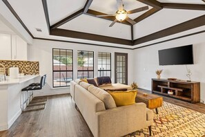 A modern living room with a sofa, coffee table, and TV. There is a kitchen area with a countertop and cabinets.