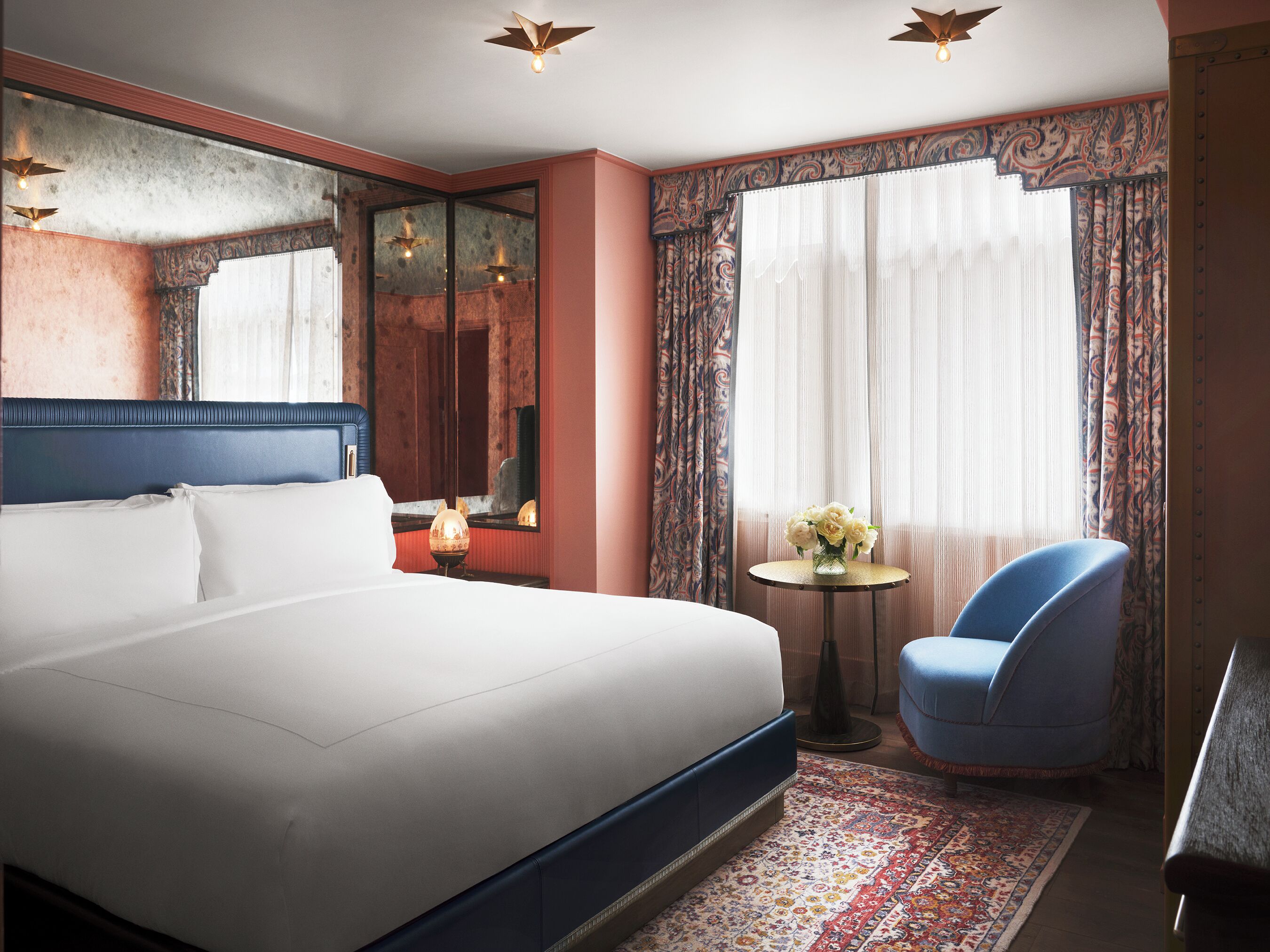 Una habitación de hotel con una cama grande, una mesita redonda con un florero, una silla y un espejo.