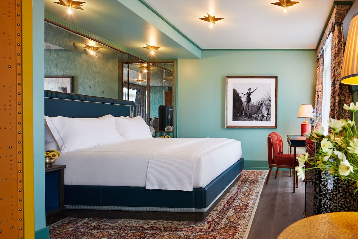 A neatly made bed with a blue headboard, a large mirror, and a framed picture on the wall.