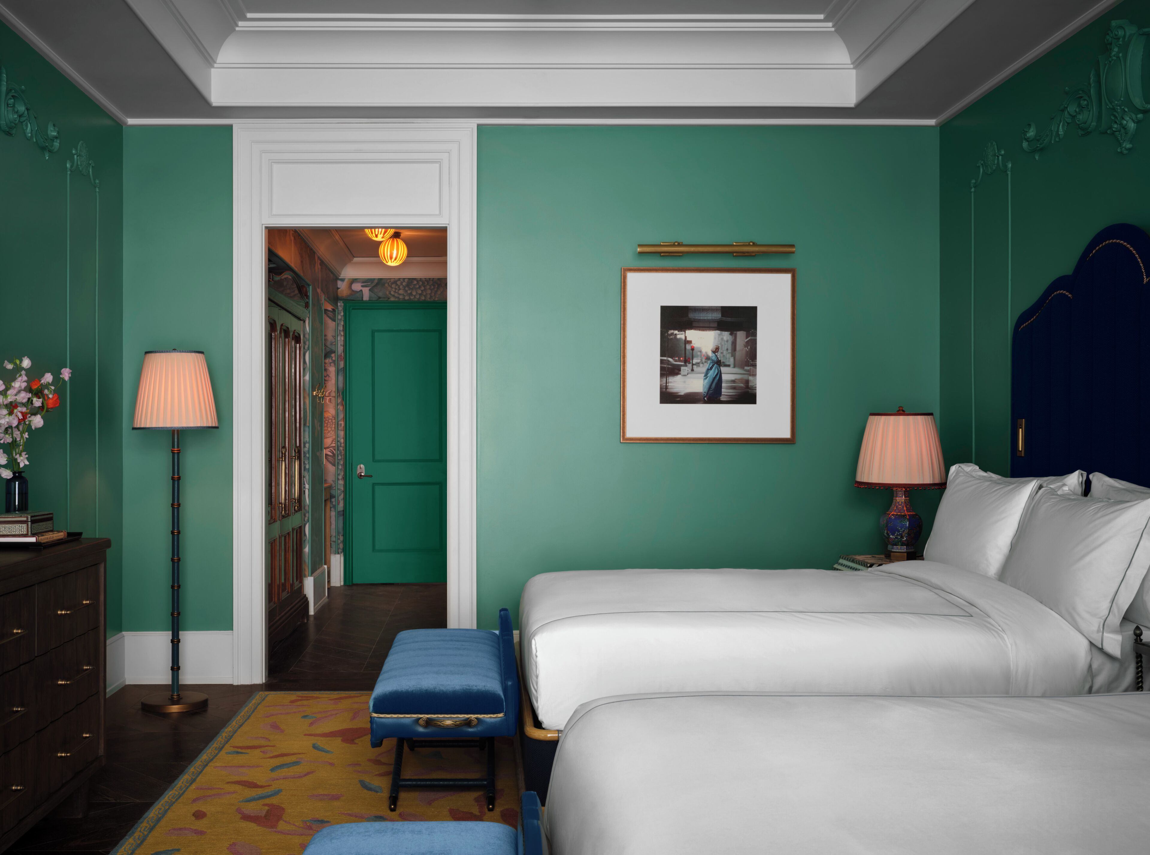 A hotel room with two beds, a desk, a chair, a lamp, a picture on the wall, and a door leading to another room.
