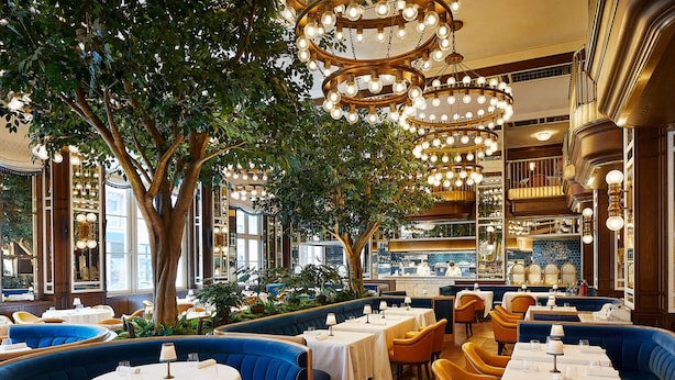 A restaurant with round tables, blue upholstered booths, and a bar area. - The Fifth Avenue Hotel (New York)