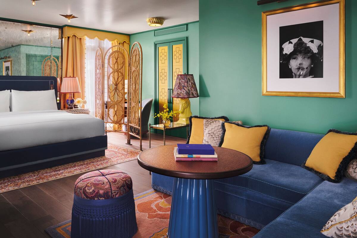 A bedroom with a bed, a blue sofa, a round table with books, a patterned ottoman, and a decorative lamp.
