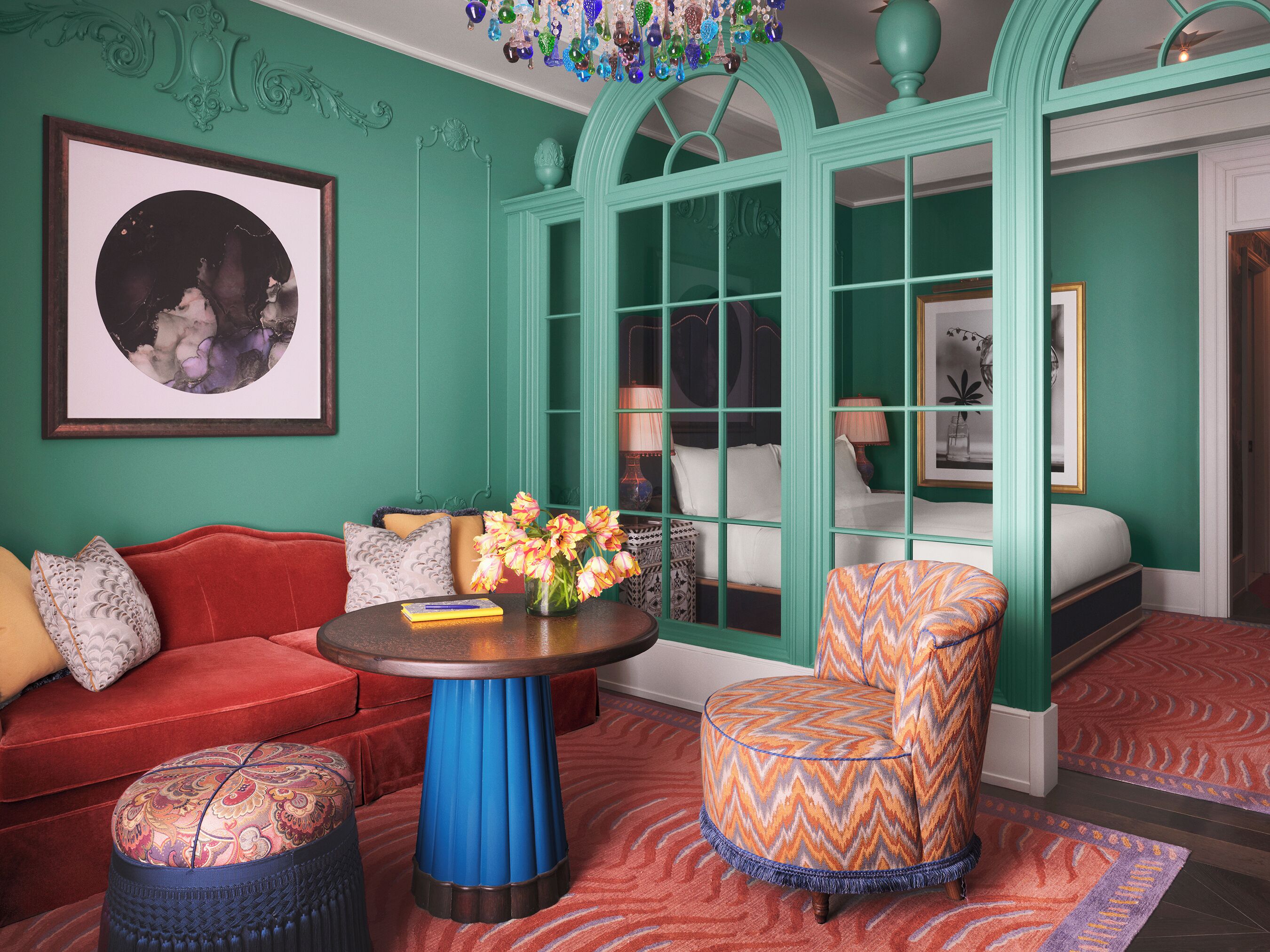Una habitación con un sofá rojo, un reposapiés estampado, una mesa redonda con un florero, un sillón estampado y una cama con pared verde y ventana.