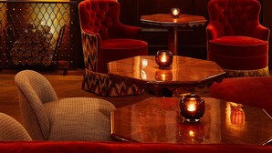 A warmly lit lounge area with a fireplace, red upholstered chairs, a round coffee table, and various framed artworks on the walls. - The Fifth Avenue Hotel (New York)
