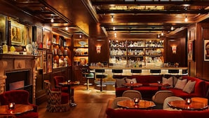 A bar area with wooden paneling, a fireplace, and a bar counter with bottles. There are red upholstered chairs and tables with candles. - The Fifth Avenue Hotel (New York)