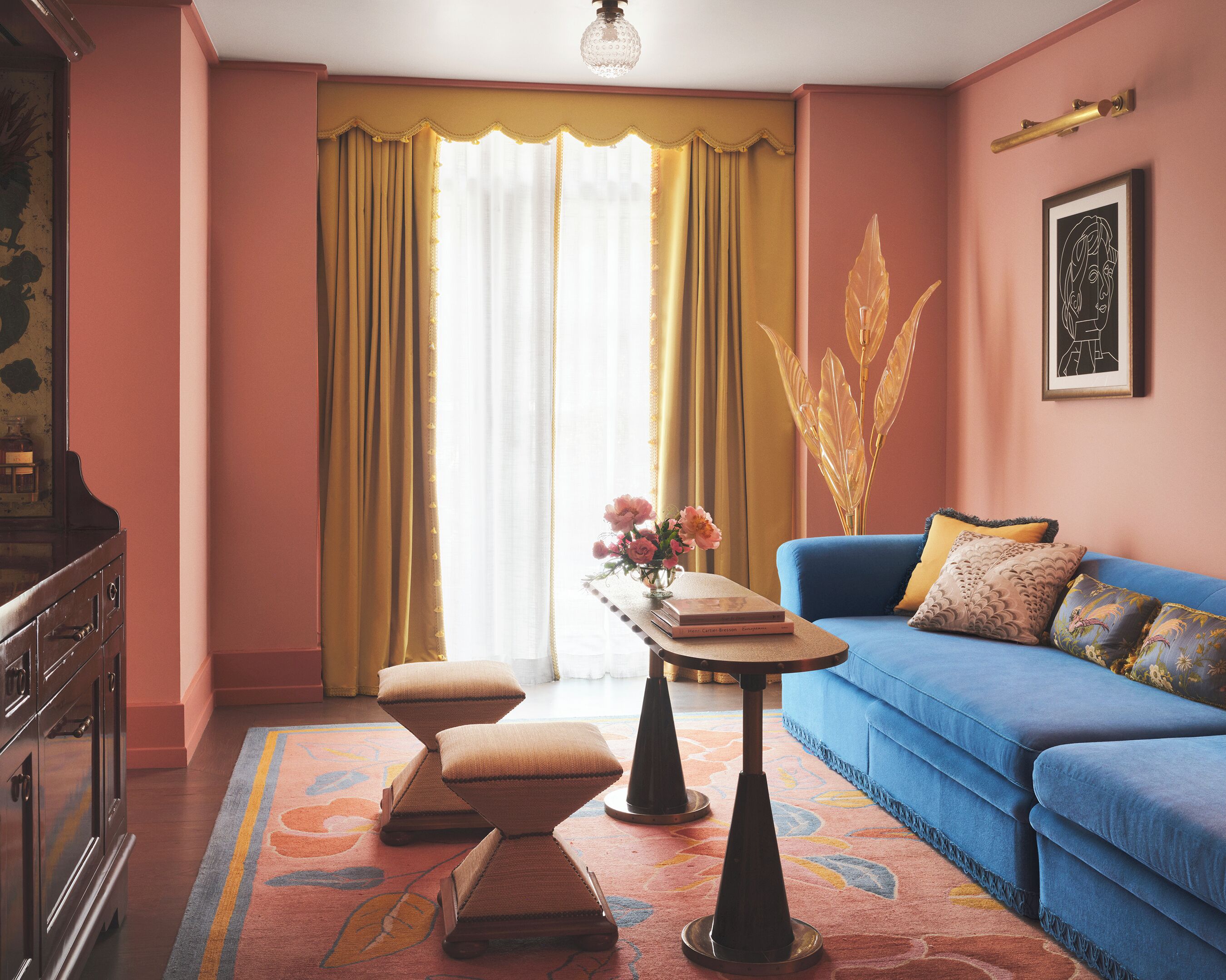 A room with a blue sofa, a wooden cabinet, a table with a vase of flowers, and a large window with curtains.