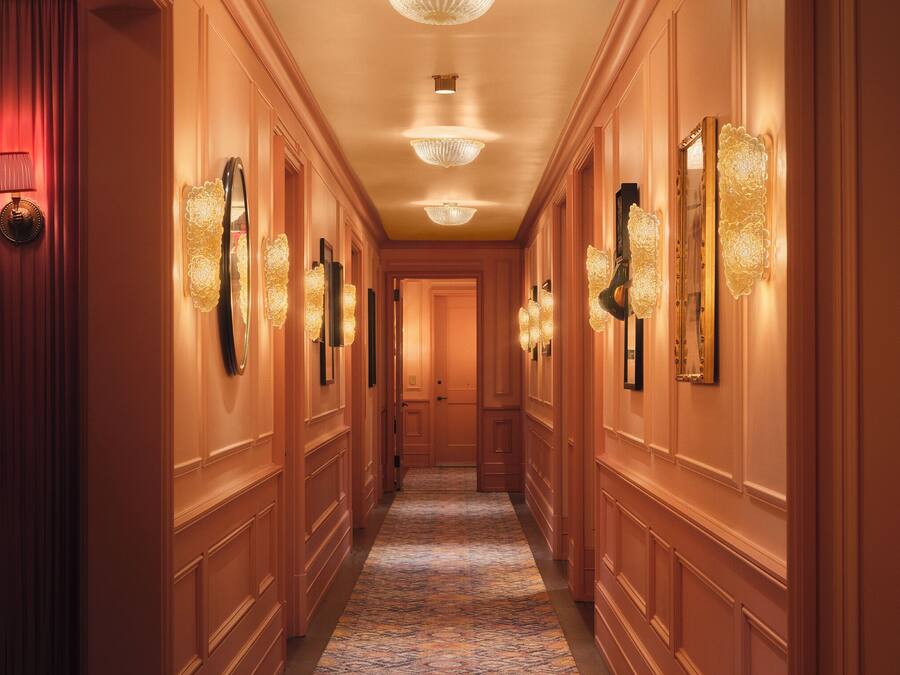 A hotel hallway with ornate wall sconces, a patterned carpet, and a polished wooden floor.