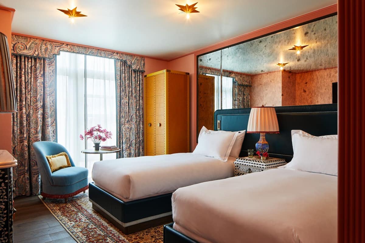 A hotel room with two beds, a chair, a small table with a lamp, and a large mirror.