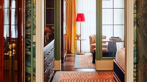 A hallway with a patterned carpet, a wooden door, and a room with a red lamp and a chair. - The Fifth Avenue Hotel (New York)