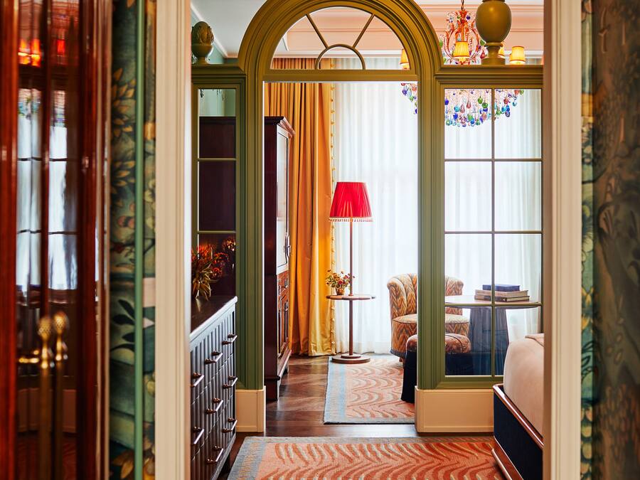 A hallway with a patterned carpet, a wooden door, and a room with a red lamp and a chair.