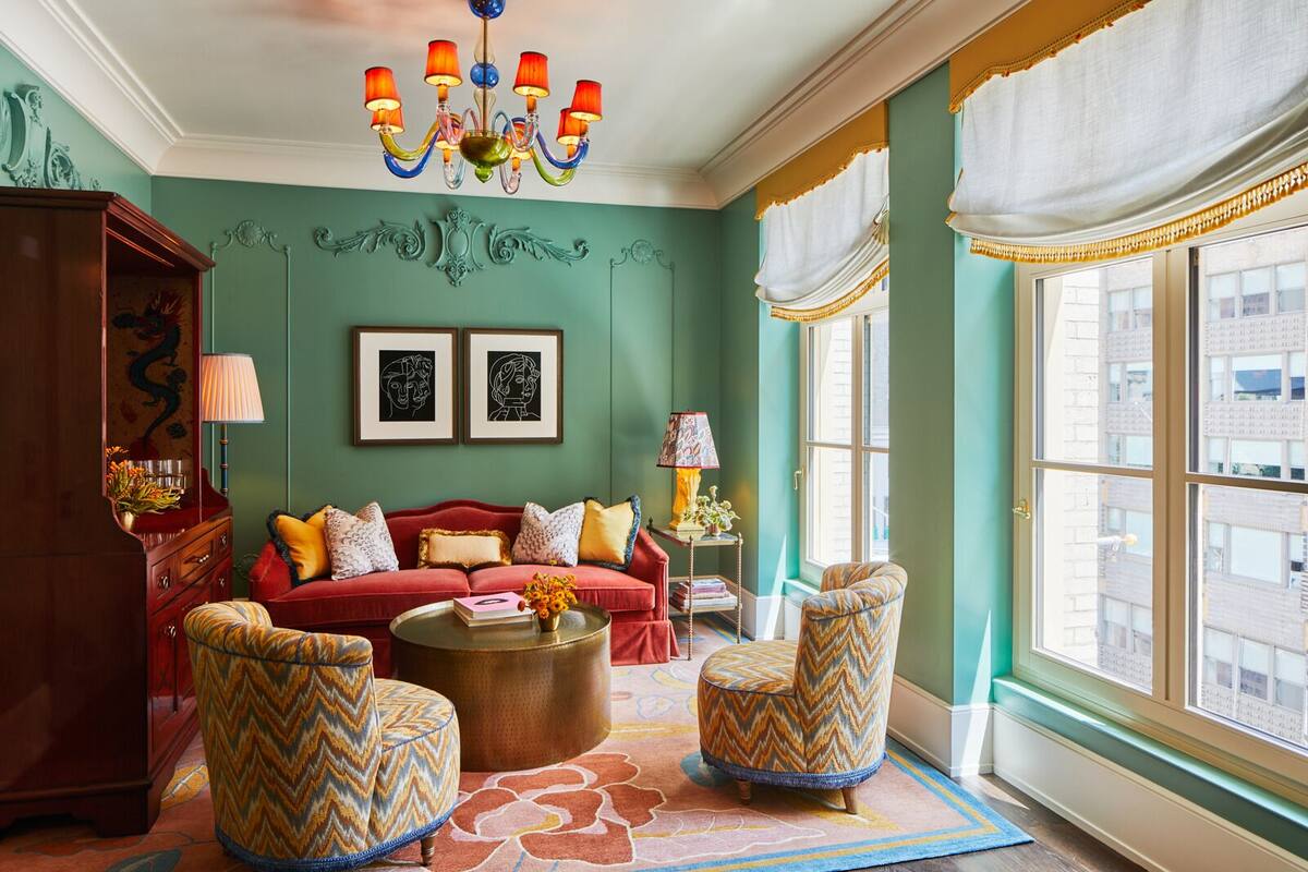 A room with a red sofa, patterned armchairs, a chandelier, framed artwork, and a large window.
