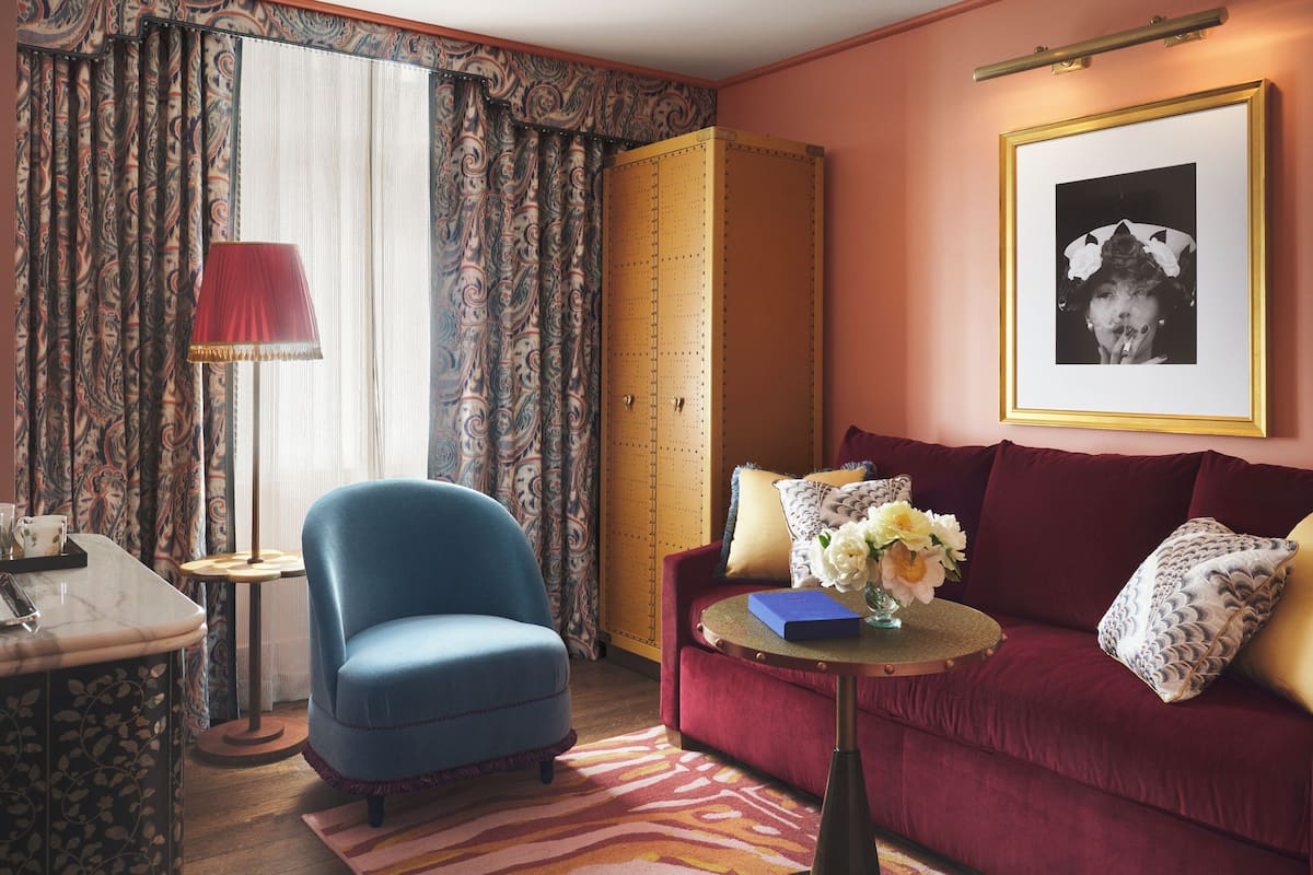 A room with a blue armchair, a red sofa, a small round table with a vase of flowers, and a framed picture on the wall.