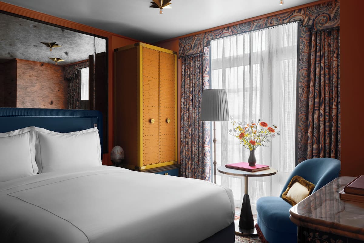 A hotel room with a large bed, a wardrobe, a small table with a vase of flowers, a chair, and a window with curtains.