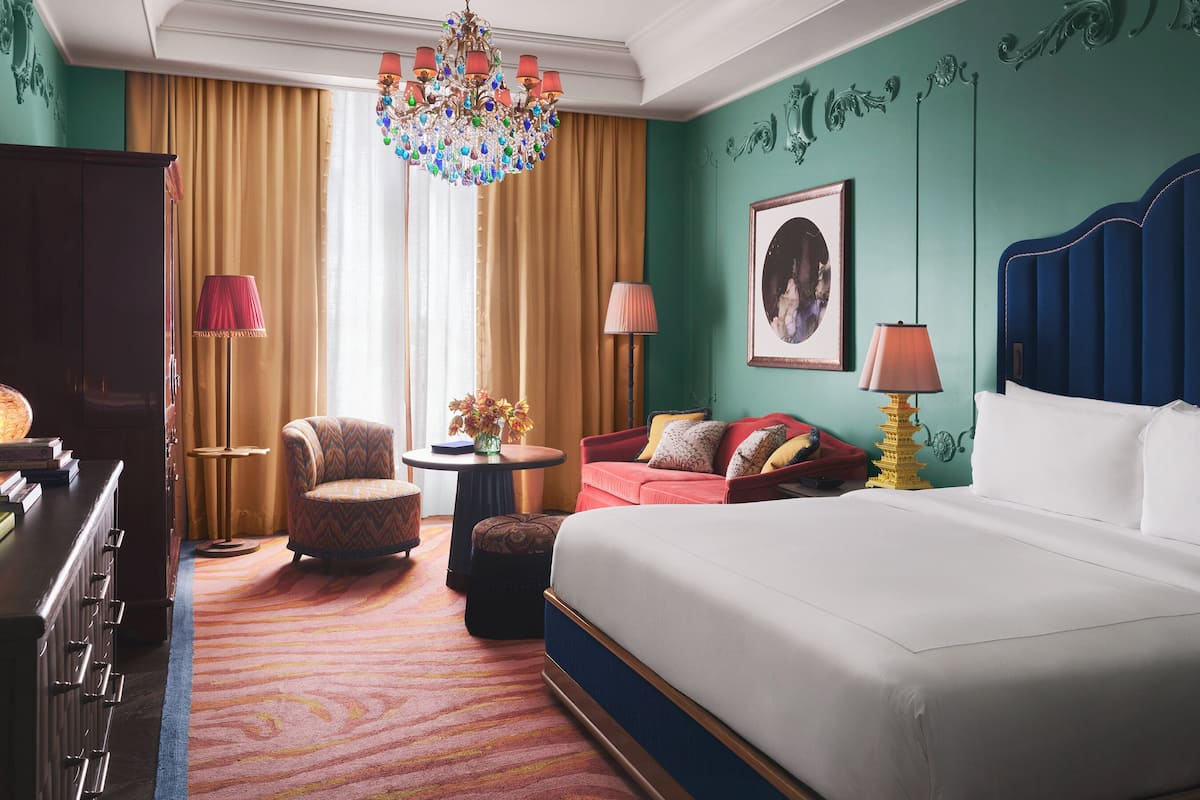 A hotel room with a large bed, a red sofa, a round table, a chandelier, and a large window with curtains.