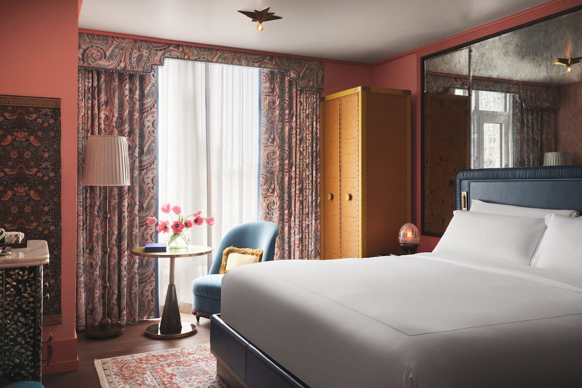 A hotel room with a bed, a chair, a table with flowers, a lamp, and a wardrobe.