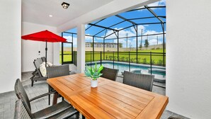 A patio with a wooden table and wicker chairs, a red umbrella, and a view of a swimming pool.