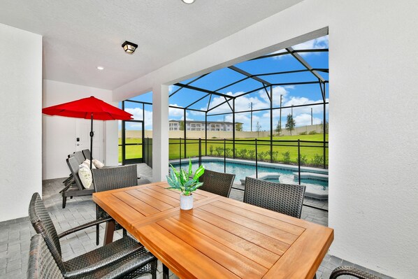 A patio with a wooden table and wicker chairs, a red umbrella, and a view of a swimming pool. - Best Value Disney Pool/Spa Home! No Carpet! No back neighbor! Resort Amenities! (Davenport)