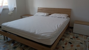 A neatly made bed with white bedding, a wooden headboard, and a nightstand with a drawer. The room has a tiled floor with a geometric pattern. - Renovated apartment in central area (Oristano)