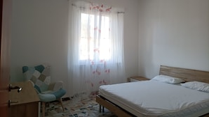 A bedroom with a bed, a chair, a nightstand, and a window with sheer curtains. - Renovated apartment in central area (Oristano)