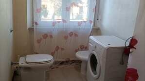 A small bathroom with a toilet, washing machine, and a window with floral curtains. - Renovated apartment in central area (Oristano)